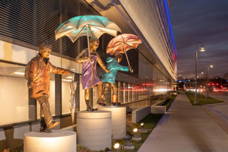Three bronze children holding umbrellas. A small Boy in an orange raincoat holds a closed umbrella, the african american girl in the middle wearing a purple jumper holind an umbrella above her head. The boy on the end wearing a turquoise rain coat and orange rain boots is holding a copper umbrella. Imagine Children's Hospital and Medical Center in Omaha, Nebraska.